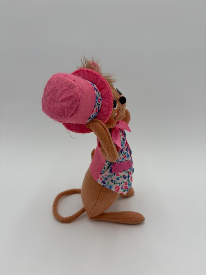May include: A brown plush mouse wearing a pink top hat, a floral vest, and a pink bow. The mouse is holding the hat, with its ears and whiskers visible. The toy is set against a white background.