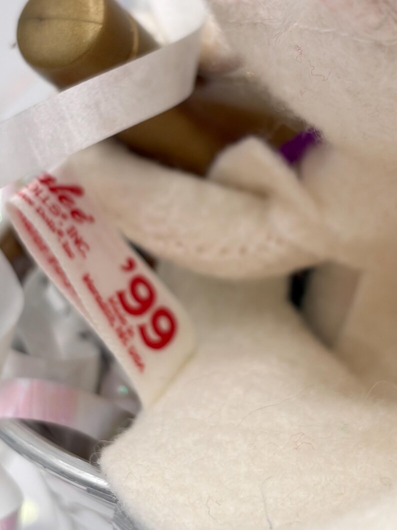 May include: Close-up of a white plush toy with a tag that reads "Lee Dolls, Inc." and the number "99" in red. The toy is partially visible, with a soft, textured appearance. White ribbon and a gold object are also in the image.