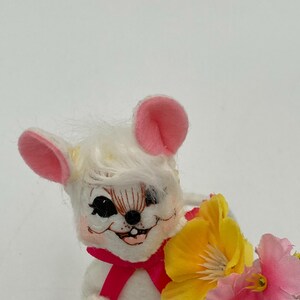 K&ouml;nnte beinhalten: Eine wei&szlig;e Pl&uuml;schmaus mit rosa Ohren und einer rosa Schleife, die einen Strau&szlig; gelber und rosa Blumen h&auml;lt. Die Maus hat ein bemaltes Gesicht mit schwarzen Augen und einem zahnigen Grinsen. Die Blumen haben gr&uuml;ne Stiele und Bl&auml;tter. Die Maus ist unten mit "Lee" beschriftet.