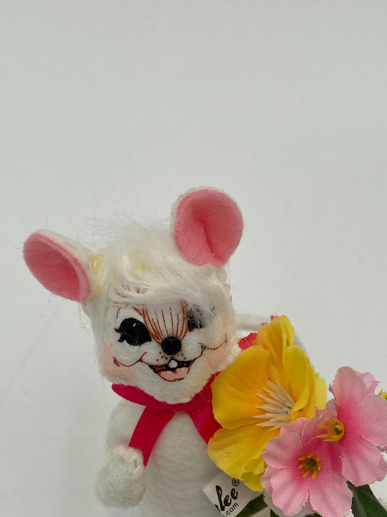 K&ouml;nnte beinhalten: Eine wei&szlig;e Pl&uuml;schmaus mit rosa Ohren und einer rosa Schleife, die einen Strau&szlig; gelber und rosa Blumen h&auml;lt. Die Maus hat ein bemaltes Gesicht mit schwarzen Augen und einem zahnigen Grinsen. Die Blumen haben gr&uuml;ne Stiele und Bl&auml;tter. Die Maus ist unten mit "Lee" beschriftet.
