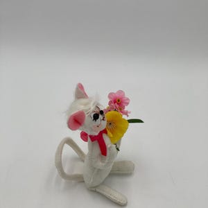 K&ouml;nnte beinhalten: Eine wei&szlig;e Filzmaus mit rosa Ohren und einer roten Schleife h&auml;lt einen Strau&szlig; rosa und gelber Blumen.