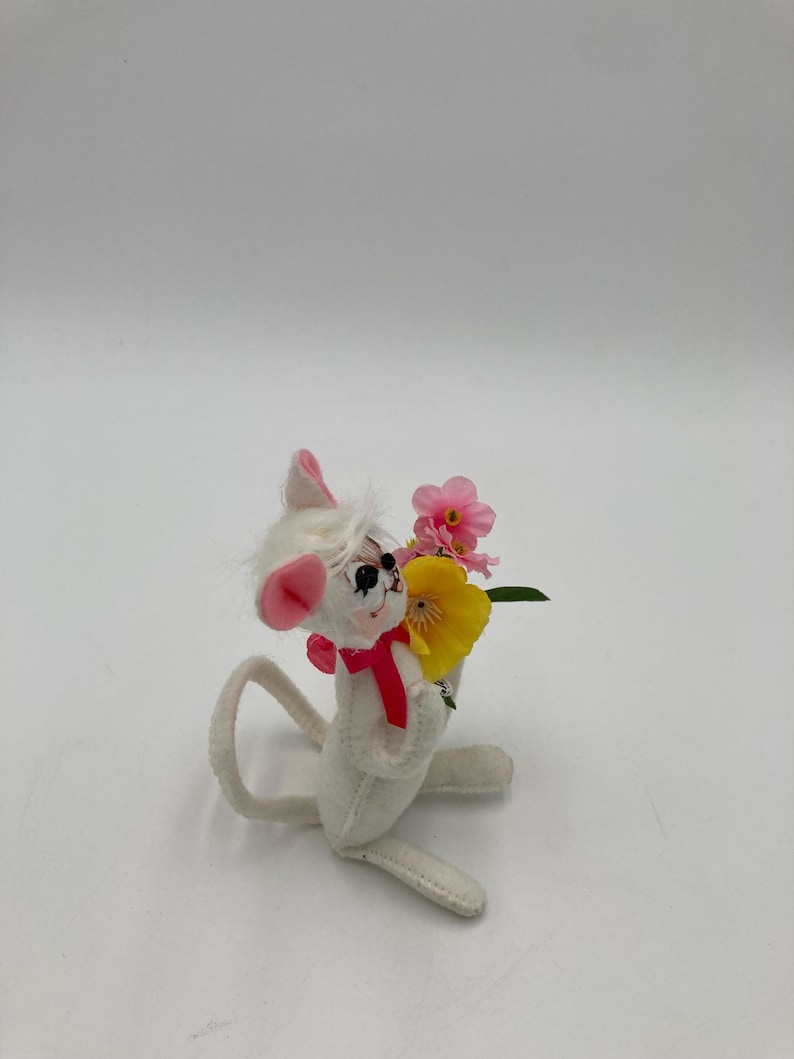 K&ouml;nnte beinhalten: Eine wei&szlig;e Filzmaus mit rosa Ohren und einer roten Schleife h&auml;lt einen Strau&szlig; rosa und gelber Blumen.