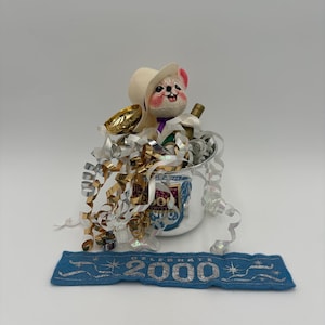 May include: A festive New Year's Eve decoration featuring a plush mouse wearing a cowboy hat, nestled in a silver bucket with a bottle of champagne and gold confetti. A blue banner below reads "2000".