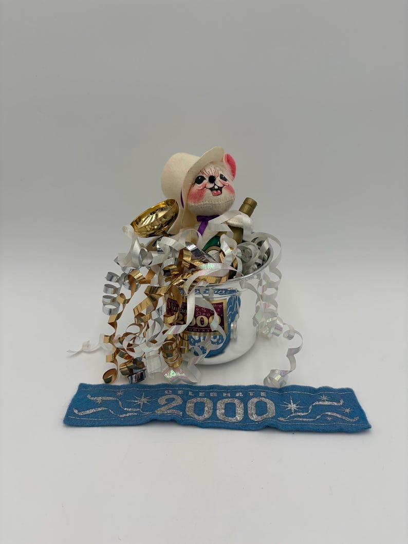 May include: A festive New Year's Eve decoration featuring a plush mouse wearing a cowboy hat, nestled in a silver bucket with a bottle of champagne and gold confetti. A blue banner below reads "2000".