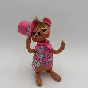 May include: A handmade felt mouse doll wearing a pink top hat and vest. The vest is pink with floral print accents and blue buttons. The mouse has brown fur, pink ears, and a pink bow tie. The doll is posed with one arm raised.
