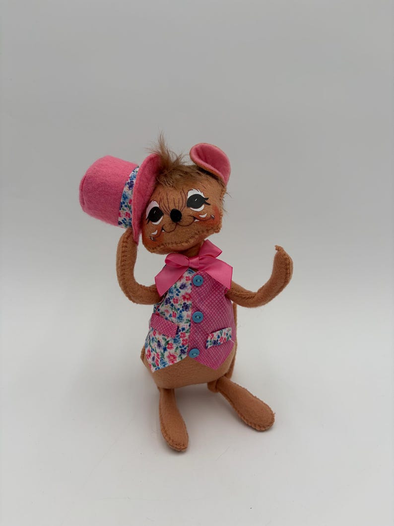 May include: A handmade felt mouse doll wearing a pink top hat and vest. The vest is pink with floral print accents and blue buttons. The mouse has brown fur, pink ears, and a pink bow tie. The doll is posed with one arm raised.