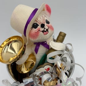 May include: A whimsical plush mouse figurine wearing a white top hat with a purple band and bow tie, holding a gold-colored trumpet and a bottle. The mouse is in a silver container with gold and silver ribbons, and a bottle of champagne.