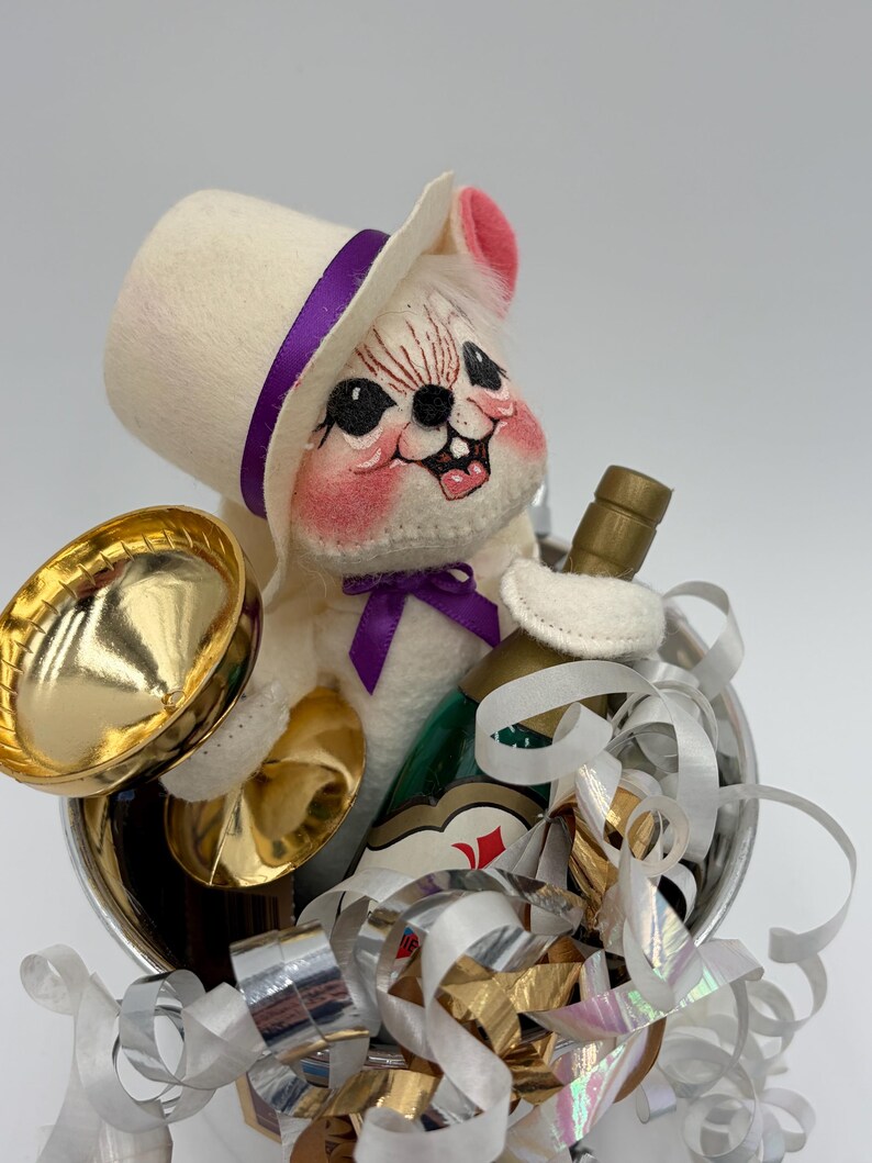 May include: A whimsical plush mouse figurine wearing a white top hat with a purple band and bow tie, holding a gold-colored trumpet and a bottle. The mouse is in a silver container with gold and silver ribbons, and a bottle of champagne.