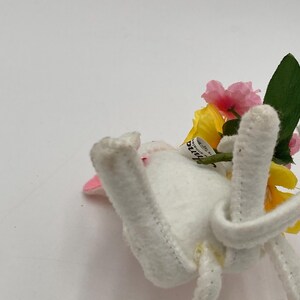 K&ouml;nnte beinhalten: Ein wei&szlig;er Filzhase mit rosa Ohren und einer gelben und rosa Bl&uuml;tenkrone. Der Hase liegt auf der Seite mit seinen Beinen in der Luft.