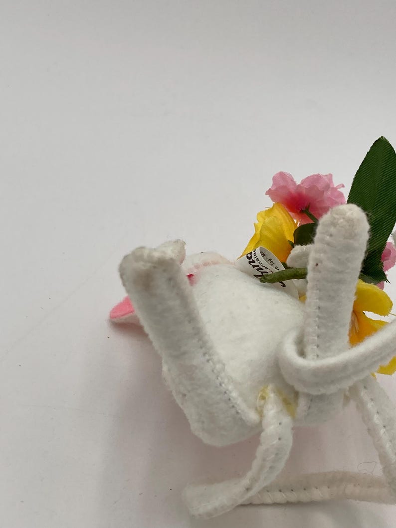 K&ouml;nnte beinhalten: Ein wei&szlig;er Filzhase mit rosa Ohren und einer gelben und rosa Bl&uuml;tenkrone. Der Hase liegt auf der Seite mit seinen Beinen in der Luft.