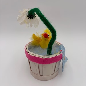 May include: A whimsical decorative piece featuring a yellow chick nestled in a small white basket with a pink rim. A large white daisy with a green stem arches over the chick. A blue and white checkered fabric is draped over the basket.
