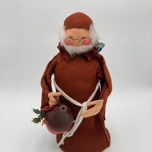 May include: A brown felt Christmas figure wearing a brown robe and a white beard, holding a brown jug with holly.