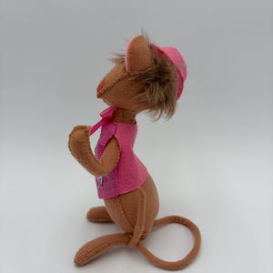 May include: A handmade brown mouse doll wearing a pink vest and hat. The mouse has brown fur and a long tail. The pink vest has a small checkered pattern. The hat has a brown fur trim and a pink ribbon.