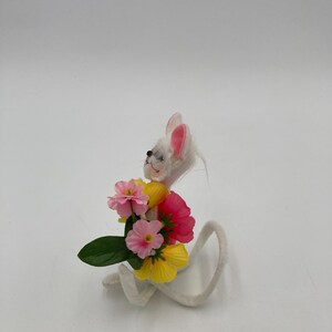 K&ouml;nnte beinhalten: Eine wei&szlig;e Filzmausfigur, die einen Strau&szlig; aus rosa, gelben und roten Filzblumen h&auml;lt. Die Maus hat rosa Ohren und einen wei&szlig;en, flauschigen Schwanz.