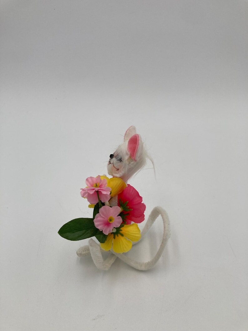 K&ouml;nnte beinhalten: Eine wei&szlig;e Filzmausfigur, die einen Strau&szlig; aus rosa, gelben und roten Filzblumen h&auml;lt. Die Maus hat rosa Ohren und einen wei&szlig;en, flauschigen Schwanz.