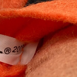 May include: Close-up of a plush toy with orange fur and a black accent. A white tag with the registered trademark symbol and the number "2000" is visible. The toy's texture appears soft and fuzzy.