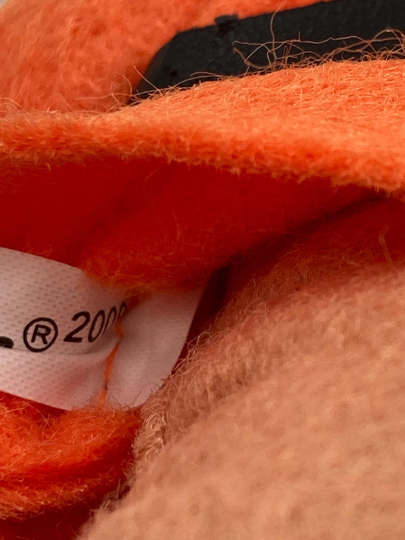 May include: Close-up of a plush toy with orange fur and a black accent. A white tag with the registered trademark symbol and the number "2000" is visible. The toy's texture appears soft and fuzzy.
