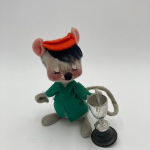 May include: A vintage stuffed mouse toy with a green jacket and an orange hat. The mouse is holding a silver trophy with a black base. The toy has gray fur and black hair.