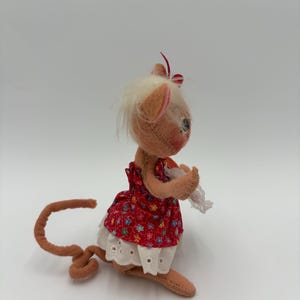 以下が含まれることがあります： 赤い花柄のドレスと白いハトメトリムを着た、手作りのぬいぐるみマウス人形。人形は、薄茶色の尻尾、耳、手足、白い髪、赤と白のストライプのリボンを持っています。白い背景に人形が置かれています。