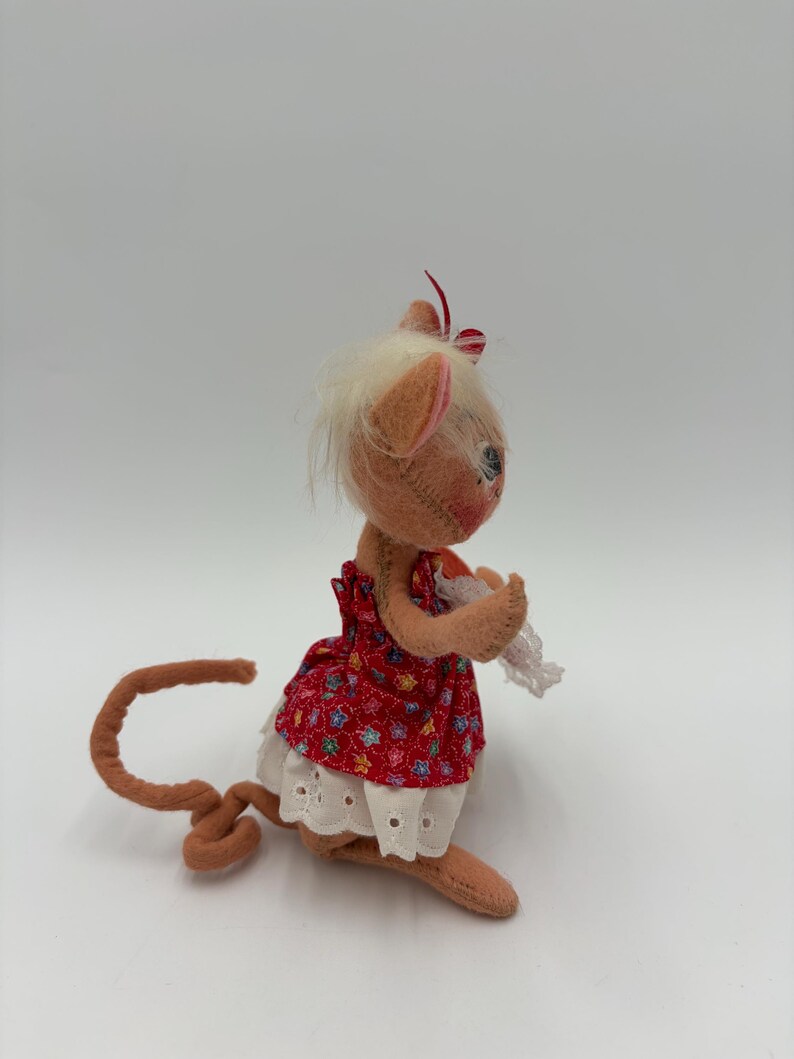 以下が含まれることがあります： 赤い花柄のドレスと白いハトメトリムを着た、手作りのぬいぐるみマウス人形。人形は、薄茶色の尻尾、耳、手足、白い髪、赤と白のストライプのリボンを持っています。白い背景に人形が置かれています。