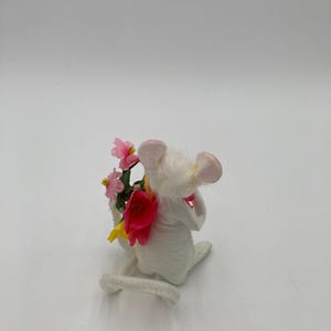 K&ouml;nnte beinhalten: Eine wei&szlig;e Filzmausfigur, die einen Strau&szlig; rosa und gelber Blumen h&auml;lt. Die Maus sitzt auf ihren Hinterbeinen, der Schwanz ist um ihren K&ouml;rper gewickelt.