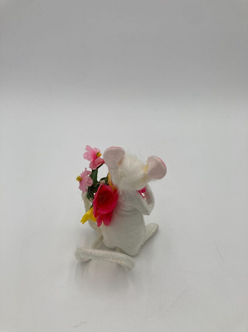 K&ouml;nnte beinhalten: Eine wei&szlig;e Filzmausfigur, die einen Strau&szlig; rosa und gelber Blumen h&auml;lt. Die Maus sitzt auf ihren Hinterbeinen, der Schwanz ist um ihren K&ouml;rper gewickelt.