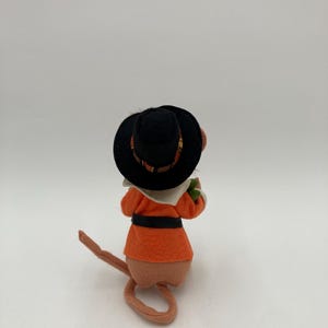 May include: A felt mouse figurine wearing an orange coat, black belt, and a black hat with a patterned band. The mouse is facing away from the camera, with its tail curled. The figurine has a tan body and a white collar.