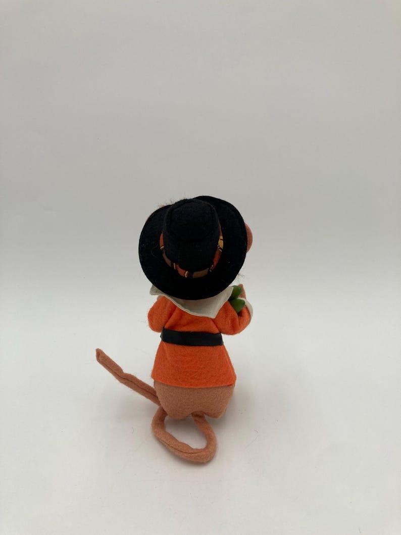May include: A felt mouse figurine wearing an orange coat, black belt, and a black hat with a patterned band. The mouse is facing away from the camera, with its tail curled. The figurine has a tan body and a white collar.