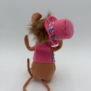 May include: A handmade plush toy mouse with brown fur, wearing a pink vest and a pink hat with a floral band. The mouse has long, thin arms and a long tail. The toy is set against a plain white background.