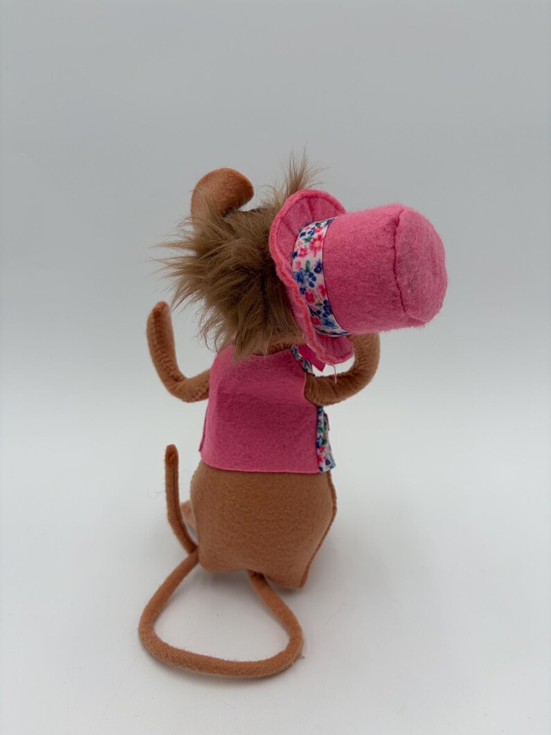 May include: A handmade plush toy mouse with brown fur, wearing a pink vest and a pink hat with a floral band. The mouse has long, thin arms and a long tail. The toy is set against a plain white background.