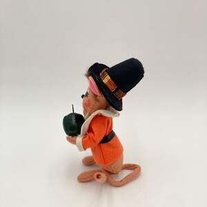 May include: A vintage figurine of a mouse dressed in a black pilgrim hat with a decorative band, an orange jacket, and a white collar. The mouse holds a dark green apple. The figurine has a long tail and is made of felt.