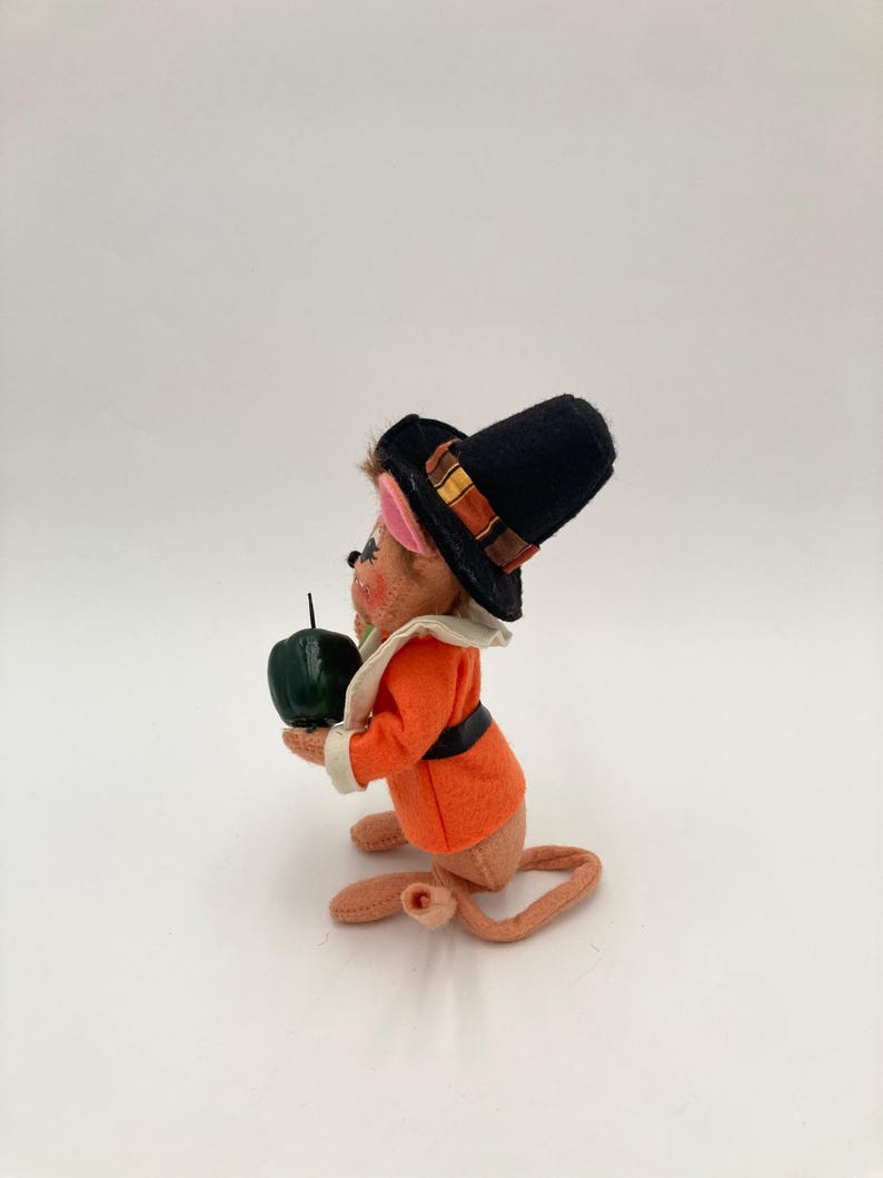 May include: A vintage figurine of a mouse dressed in a black pilgrim hat with a decorative band, an orange jacket, and a white collar. The mouse holds a dark green apple. The figurine has a long tail and is made of felt.