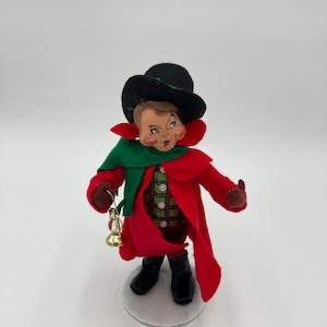 Könnte beinhalten: Eine Weihnachtswichtelpuppe aus Filz, die einen roten Mantel, einen grünen Schal, einen schwarzen Hut trägt und eine goldene Glocke hält.