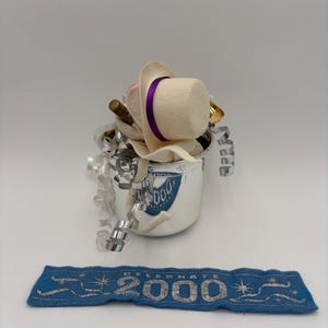 May include: A silver bucket filled with party favors, including a white hat with a purple band, a gold-colored horn, and silver streamers. A blue banner with the text "CELEBRATE 2000" is below the bucket.