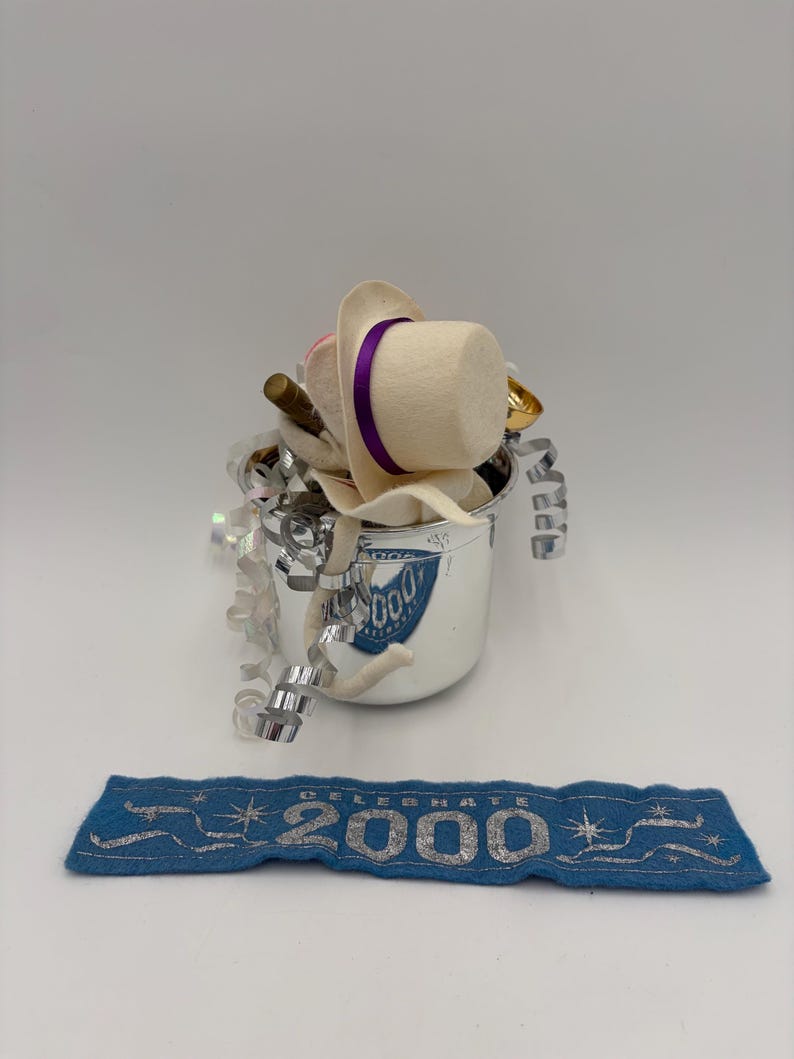 May include: A silver bucket filled with party favors, including a white hat with a purple band, a gold-colored horn, and silver streamers. A blue banner with the text "CELEBRATE 2000" is below the bucket.