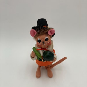 May include: A vintage felt mouse figurine dressed as a pilgrim, wearing a black hat with a brown band and an orange belt. The mouse holds a green vegetable and a dark green round object. The mouse has pink ears and a brown tail.