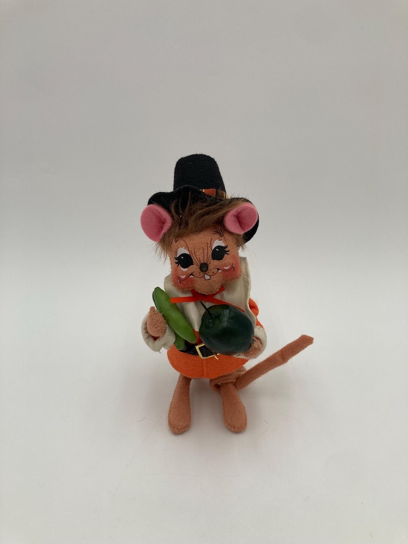 May include: A vintage felt mouse figurine dressed as a pilgrim, wearing a black hat with a brown band and an orange belt. The mouse holds a green vegetable and a dark green round object. The mouse has pink ears and a brown tail.