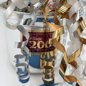 May include: A silver-colored container with gold and silver metallic streamers. The container has a label that reads "Vintage 2000". A blue fabric is visible in the foreground. The streamers are curled and add a festive touch.