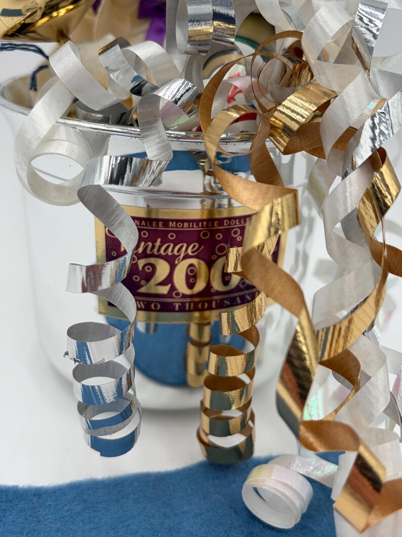 May include: A silver-colored container with gold and silver metallic streamers. The container has a label that reads "Vintage 2000". A blue fabric is visible in the foreground. The streamers are curled and add a festive touch.