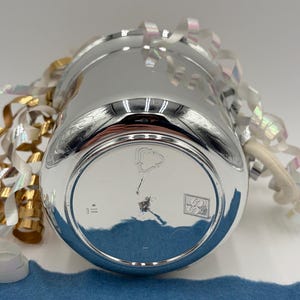 May include: A shiny, silver-colored metal cup with a rounded base. The cup is surrounded by decorative ribbons in gold, white, and iridescent colors. A blue felt strip is visible in the foreground.