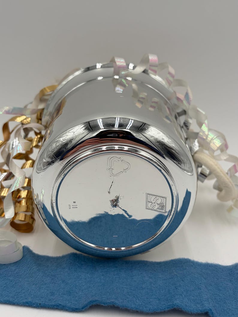 May include: A shiny, silver-colored metal cup with a rounded base. The cup is surrounded by decorative ribbons in gold, white, and iridescent colors. A blue felt strip is visible in the foreground.