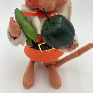 May include: A vintage felt mouse figurine wearing an orange outfit with a black belt and a black hat. The mouse holds a green vegetable and a dark green fruit. The figurine has a tan body and face with a painted expression.