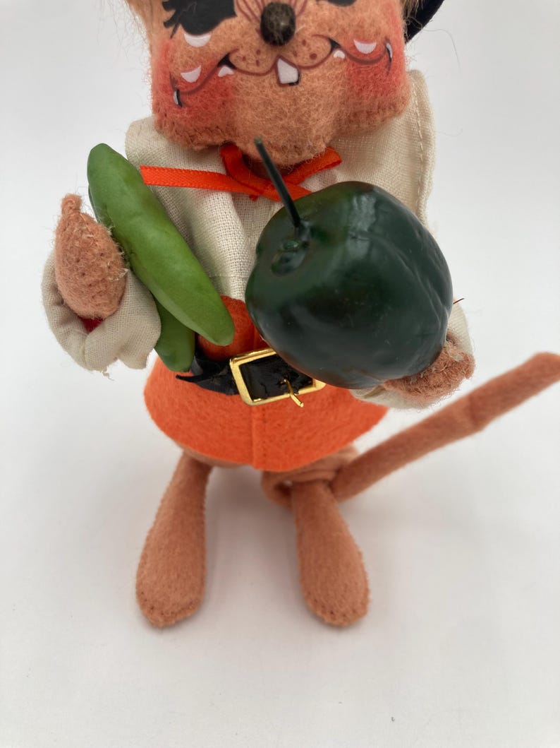 May include: A vintage felt mouse figurine wearing an orange outfit with a black belt and a black hat. The mouse holds a green vegetable and a dark green fruit. The figurine has a tan body and face with a painted expression.