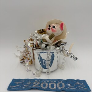 May include: A vintage silver-colored cup with a blue emblem, filled with metallic streamers and a small stuffed animal wearing a hat. A blue felt banner below reads "2000" in silver lettering, celebrating the new millennium.