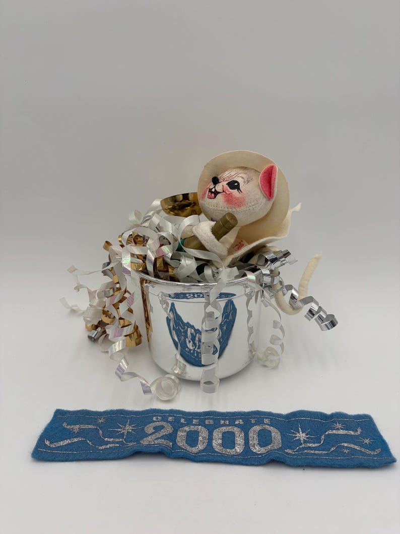 May include: A vintage silver-colored cup with a blue emblem, filled with metallic streamers and a small stuffed animal wearing a hat. A blue felt banner below reads "2000" in silver lettering, celebrating the new millennium.