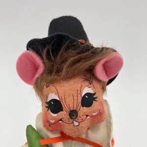May include: A vintage-style plush mouse figurine wearing a black hat with an orange band, a white outfit with an orange ribbon, and holding green vegetables. The mouse has pink ears, brown hair, and a smiling face.