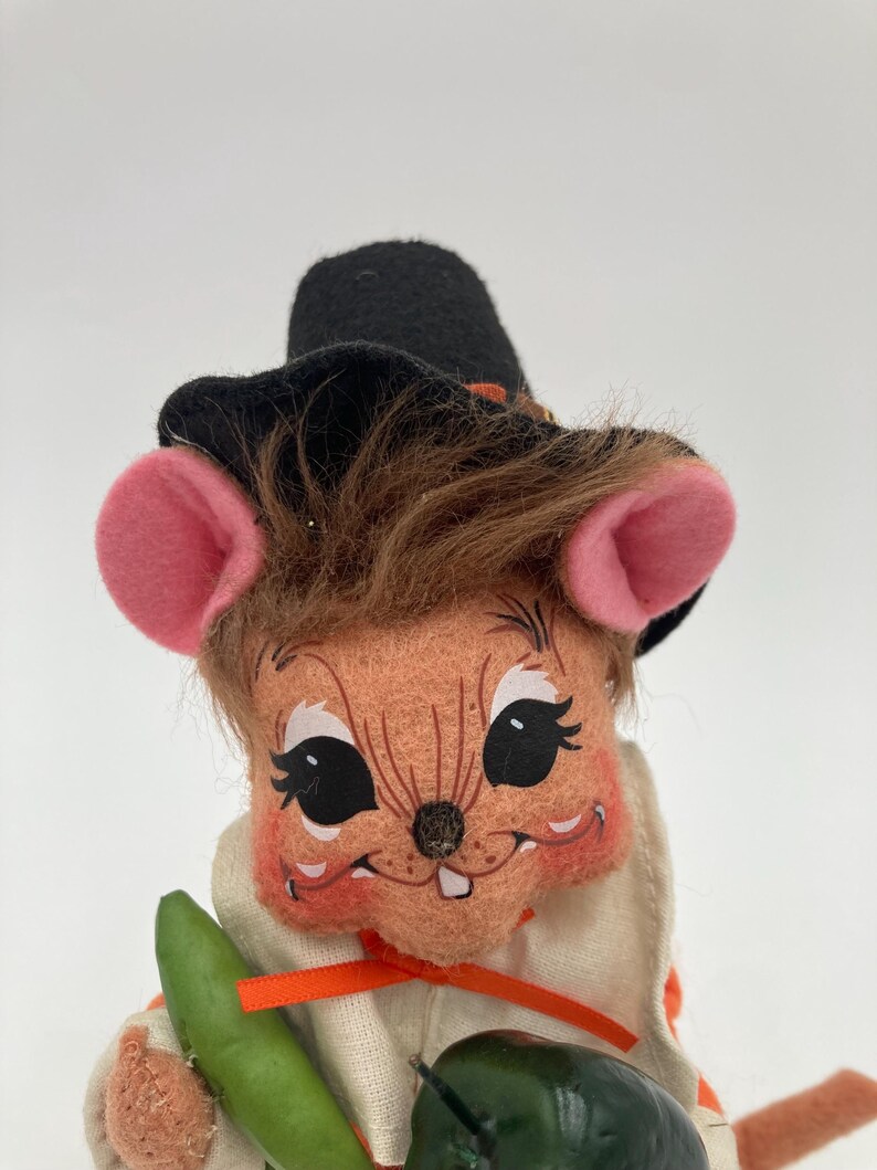 May include: A vintage-style plush mouse figurine wearing a black hat with an orange band, a white outfit with an orange ribbon, and holding green vegetables. The mouse has pink ears, brown hair, and a smiling face.