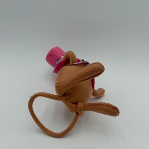 May include: A brown felt bunny toy with long ears and a pink hat with a floral pattern. The bunny is lying on its back, with its arms and legs extended. The hat has a pink brim and a floral band.