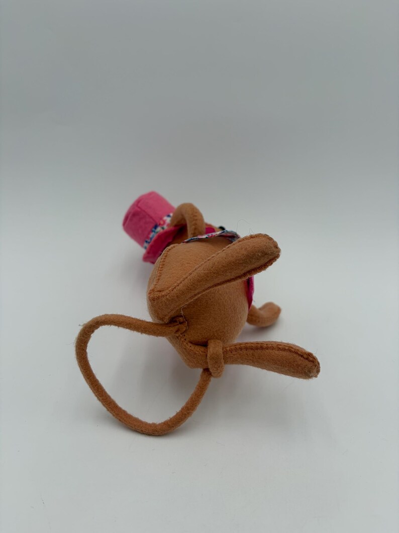 May include: A brown felt bunny toy with long ears and a pink hat with a floral pattern. The bunny is lying on its back, with its arms and legs extended. The hat has a pink brim and a floral band.