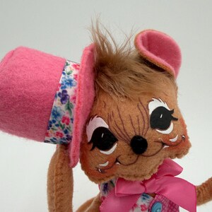 May include: A handmade plush toy mouse with a pink top hat and a floral band. The mouse has brown fur, large black eyes, and a pink bow. The mouse is wearing a pink vest with blue buttons and a floral print dress.